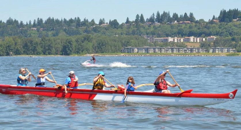 Six paddlers of various ages and sizes are on a red and white outrigger canoe on the Columbia River. There is a jet ski on the water behind the canoe, and Vancouver, Washington is visible in the background. 